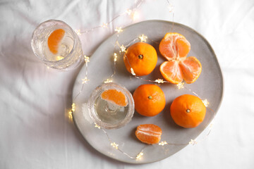 Delicious ripe tangerines, glasses of sparkling wine and fairy lights on white bedsheet, flat lay
