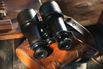 Vintage military binoculars in the leather case, laying on the brown leather case on the wooden brown table.
