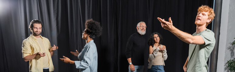 young redhead man posing with outstretched hand near interracial actors talking in acting school, banner.