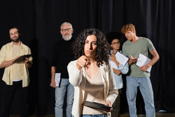 multiracial woman with serious face expression pointing at camera while rehearsing near blurred actors and mature art director.