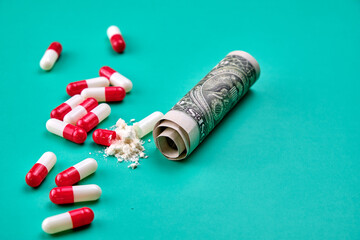 Red and white capsules, one of them is open, you can see the content that is a white powder, next to it there is a rolled dollar bill on a green table.