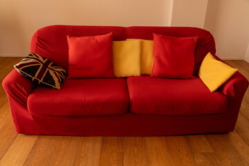 on a parquet floor three seater sofa upholstered in red fabric and trimmed with two red cushions, three yellow cushions and an English cushion