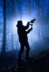 Silhouette of a videographer in a misty forest, dramatically backlit with cool blue color and rays...