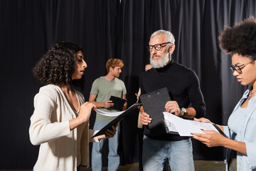bearded acting skills teacher looking at interracial students holding clipboards with screenplays.