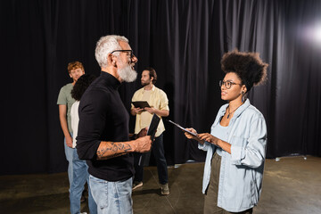 tattooed grey haired screenwriter holding scenario near african american woman with clipboard and...