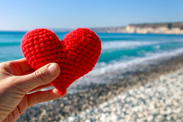 Close-up of red crochet heart in hand on sea beach - obrazy, fototapety, plakaty