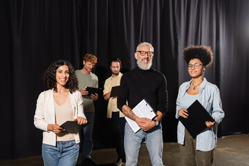bearded and tattooed art director with interracial actresses smiling at camera in acting studio.