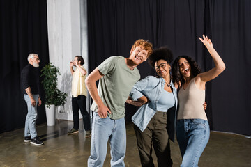 excited multiracial actress waving hand near happy interracial team in theater.