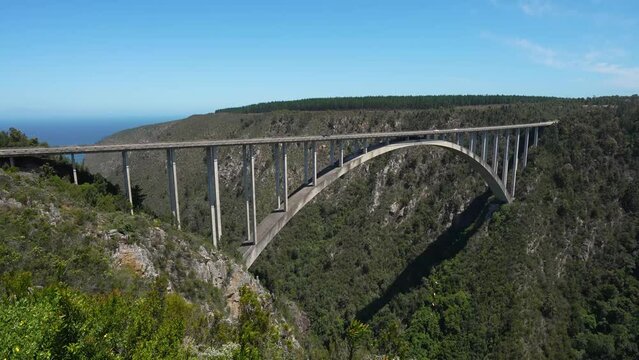 4K video of cars and trucks passing over an arch bridge closing the gap over a lush green jungle bush valley for cars and truck to safely travel to their destination. Midday sun baking the landscape