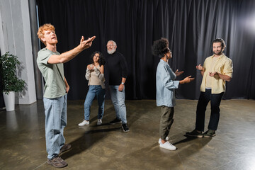 redhead man rehearsing with outstretched hand near art director and multiethnic actors talking on stage in theater.