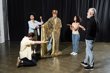 redhead man in king costume rehearsing role near multiethnic actors and art director in acting school.