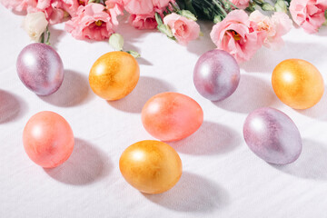 Obraz na płótnie Canvas Gold, purple and pink painted Easter eggs in pastel colors and flower bouquet on a white cotton tablecloth