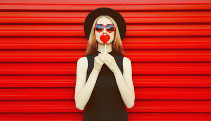 Portrait of beautiful young woman with lollipop blowing her lips with lipstick sending sweet air kiss wearing red heart shaped sunglasses, black round hat on background