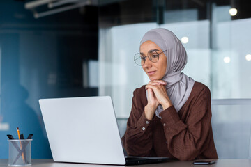 Successful smiling Arab woman in hijab working inside modern office, Muslim woman using laptop at...