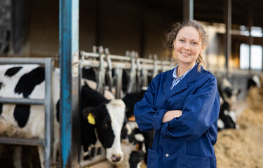 Positive woman farmer standing in cowshed with arms crossed and smiling.