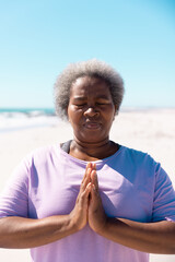 African american senior woman with eyes closed meditating in prayer pose at beach under clear sky