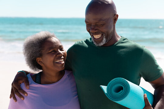 Smiling african american senior couple with exercise mat standing against sea and sky on sunny day - Powered by Adobe