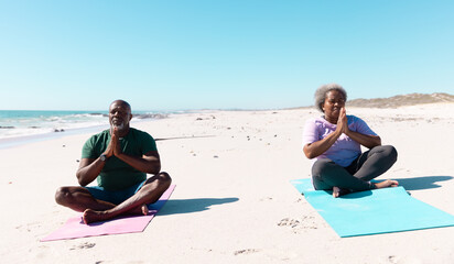 African american senior couple with cross legged sitting and meditating on beach against clear sky