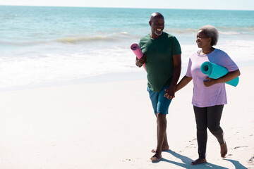 Cheerful african american senior couple with mats holding hands while walking at beach on sunny day