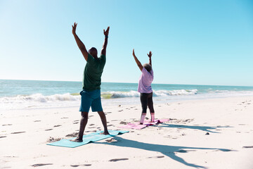 African american senior couple with arms raised exercising on mats at beach under clear blue sk