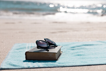 Close-up of book with sunglasses and blue towel on sandy beach against sea during sunny day