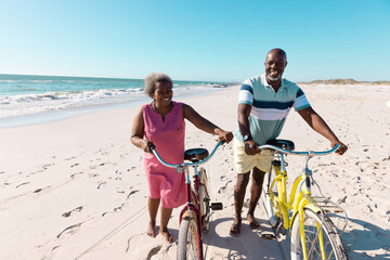 African american senior couple with bicycles walking at sandy beach under clear sky during vacation