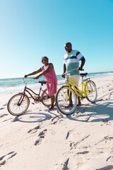 Cheerful african american senior couple with bicycles talking and walking at beach under clear sky
