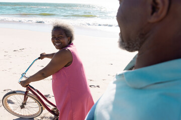 Happy african american senior woman with bicycle walking at beach and talking with man on sunny day