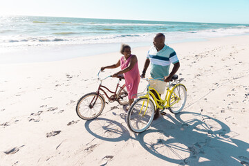 High angle view of african american senior couple with bikes talking and walking at beach in summer