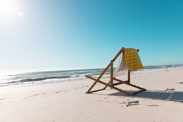 Empty deckchair with yellow napkin on sandy beach under clear blue sky during sunny day - obrazy, fototapety, plakaty