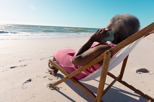 African American Senior Woman Talking Over Mobile Phone While Relaxing On Deckchair At Beach