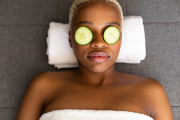 Happy african american woman lying on towel with cucumber slices on her eyes