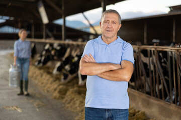 Portrait of cheerful middle-aged man farmer standing in cowhouse with arms crossed. Confident man dairy farm worker.