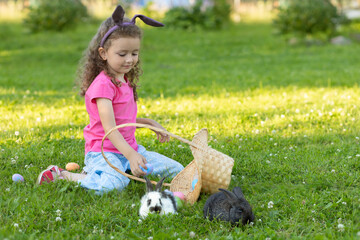 Fototapeta na wymiar little cute girl with hear ears playing with two rabbits in basket in green grass. Child with pet bunny outdoors. Happy kid with animal. Children at easter egg hunt