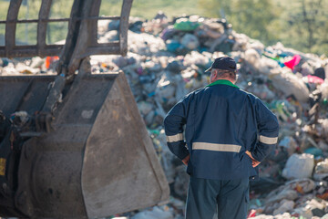Man in a blue working suit looking at garbage near landfill skid steer loader