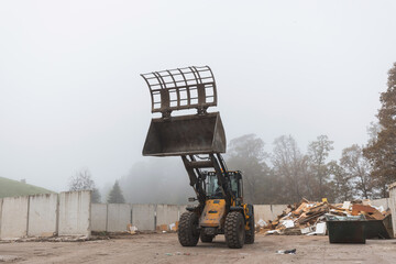 Obraz premium Yellow wheel loader, with lifted scrap grapple, moving along the recycling center area in process handling dumped waste, handheld shot.
