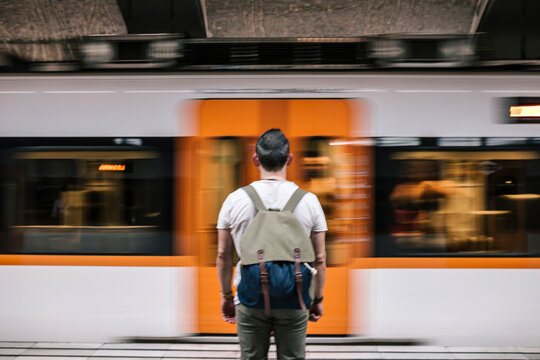 Rear View Of Man With Backpack Looking At Moving Train While Standing On Railroad Station Platform