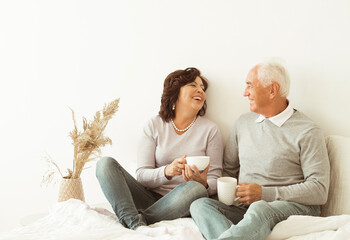 Happy elderly couple drinking coffee sitting on bed on bedroom.