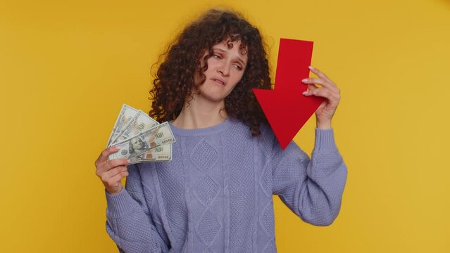 Upset curly haired woman showing red arrow pointing down, concept of downgrade, unsuccessful business, fall of stock market money dollar exchange rate bankruptcy fail. Young girl on yellow background