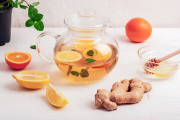 Glass teapot with orange, lemon, ginger and mint tea with ingredients on white wooden table