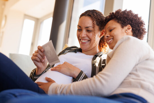Happy diverse lesbian couple sitting on sofa, looking at usg scan and embracing