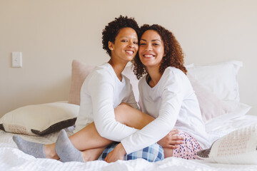 Portrait of happy diverse lesbian couple sitting on bed and embracing