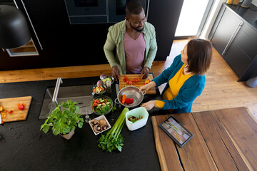 High angle of happy diverse couple preparing food and using tablet in kitchen, copy space