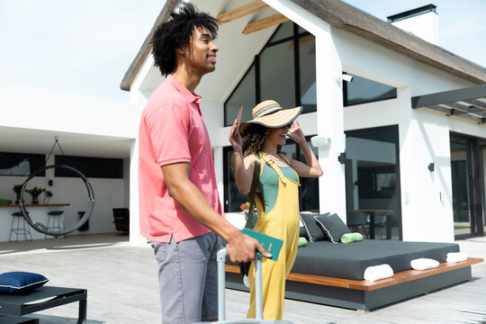 Happy biracial couple arriving outside holiday home with suitcase, copy space