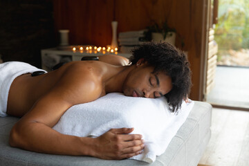 Relaxed biracial man on massage table having hot stones treatment at spa, copy space