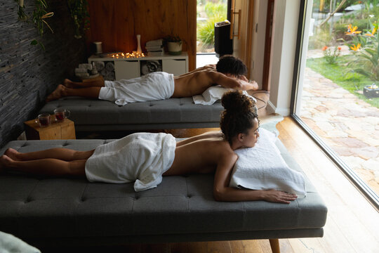 Relaxed biracial couple wearing towels lying on massage tables at health spa, copy space