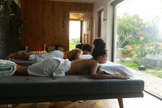 Relaxed biracial couple wearing towels lying on massage tables at health spa, copy space