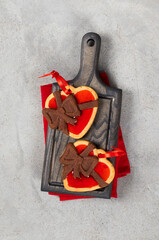 Two Cookies with a chocolate bow, heart-shaped with strawberry marmalade filling, with a red satin ribbon. On a serving board, on a red linen napkin. Light gray background. Valentine's day. Top view