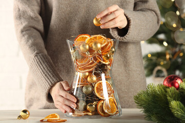 Woman putting Christmas tree ball into glass vase with dry orange slices and cones at white wooden...