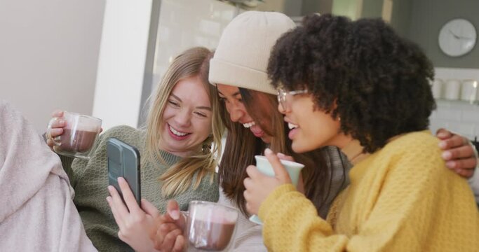 Happy diverse teenager girls friends drinking coffee and using smartphone at home, slow motion